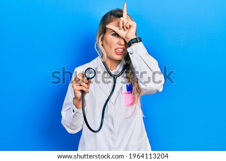 Beautiful young blonde doctor woman holding stethoscope making fun of people with fingers on forehead doing loser gesture mocking and insulting. 