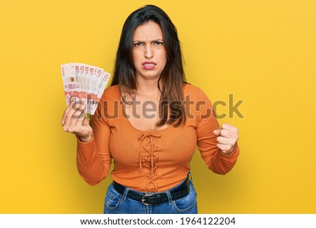 Beautiful hispanic woman holding 10 colombian pesos banknotes annoyed and frustrated shouting with anger, yelling crazy with anger and hand raised 