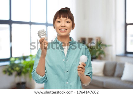 eco living, inspiration and sustainability concept - portrait of happy smiling young asian woman in turquoise shirt holding energy saving lighting bulb over home room background