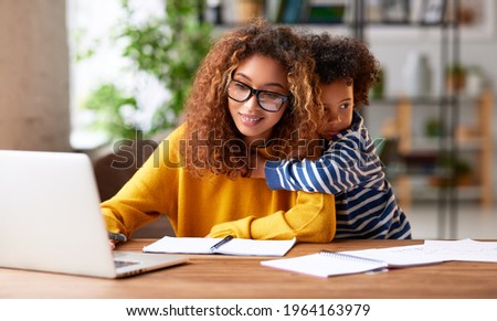 Working with kids. Young focused woman mother wearing eyeglasses using laptop and thinking about work task while small boy son gently hugs her. Childcare concept