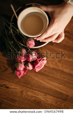 The woman's hand holds a cup of coffee, and pink roses lie next to it. Photo vertical