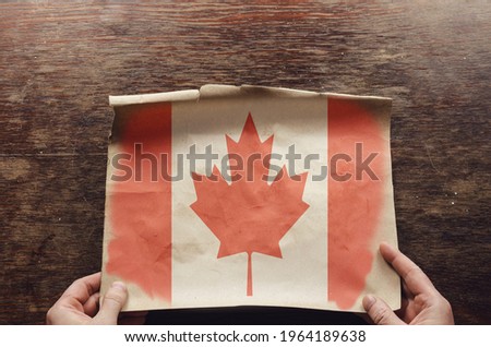 An old, shabby piece of paper with the Canadian flag on it lies on the scratched table. The man's hands hold the unfolded piece of parchment to the brown wooden table. Top view