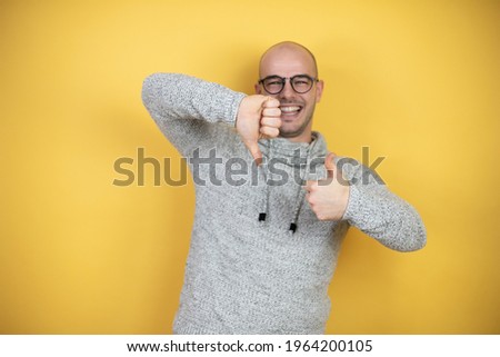 Young bald man wearing glasses over yellow background Doing thumbs up and down, disagreement and agreement expression. Crazy conflict