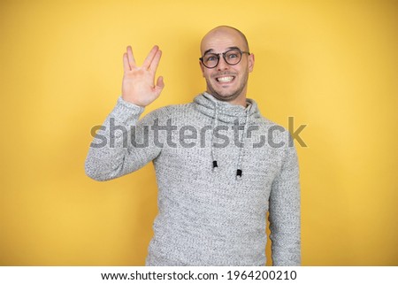 Young bald man wearing glasses over yellow background doing hand symbol