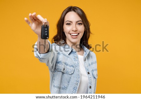 Young smiling happy satisfied excited successful brunette woman 20s in denim shirt white t-shirt hold in hands giving car keys show thumb up like gesture isolated on yellow background studio portrait Royalty-Free Stock Photo #1964211946