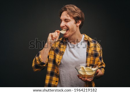 Young blond man in yellow shirt is eating chips and holding plate with it, isolated over black background Royalty-Free Stock Photo #1964219182