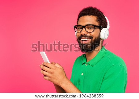 african american man in glasses and wireless headphones texting on smartphone isolated on pink