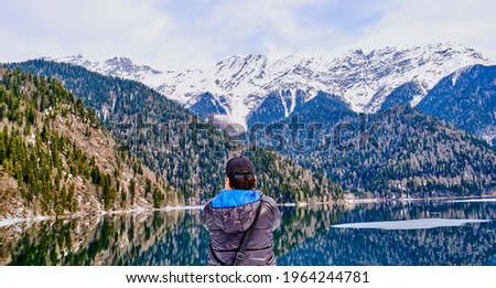 A man in a black jacket with a blue hood takes a picture of a beautiful winter landscape of the Caucasus mountains and Lake Ritsa, located in Abkhazia
