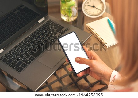 Woman using smartphone next to laptop, mockup with blank white screen, concept photo of contact business searching information in workplace 