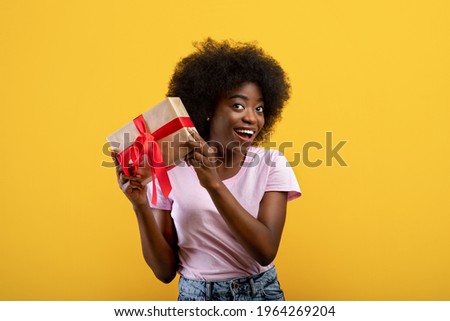 Guess what's inside. Happy black woman listening what rattles in gift box, yellow studio background. Positive lady shaking present and looking at camera, thinking and smiling