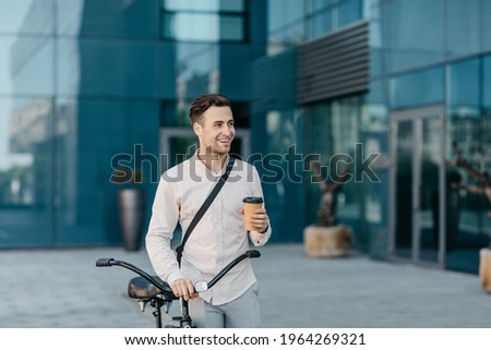 Morning coffee, road at job with eco transport and start work day. Smiling millennial handsome guy in formal wear with bicycle and cup of hot drink on office building background, outdoor, free space