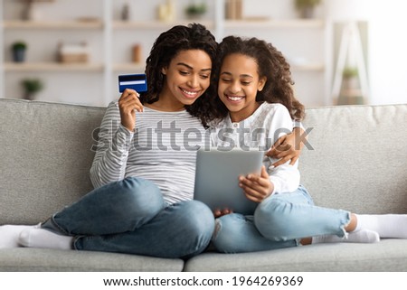 Cute african american mother and kid shopping together on digital tablet, holding credit card, sitting and hugging on couch at home, looking at pad screen and smiling, order food, copy space Royalty-Free Stock Photo #1964269369