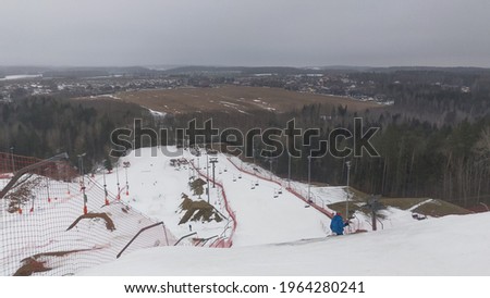 A snow-covered ski and snowboard track, equipped with a chairlift, mesh fence and light poles, surrounded by forest and with views of fields and villages in the background in early spring.
