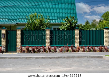 Green metal corrugated fence with brick pillars on the background of a beautiful house. Royalty-Free Stock Photo #1964317918