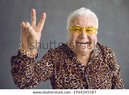 Close up of cheerful stylish glamorous senior man showing v-sign peace symbol on gray background. Pensioner in glasses, a shirt with a leopard print and a gold bracelet on his arm is smiling happily.