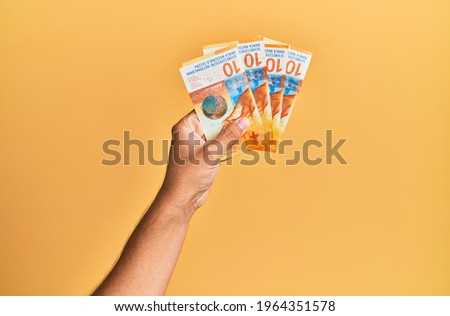 Hand of hispanic man holding swiss 10 franc banknotes over isolated yellow background.
