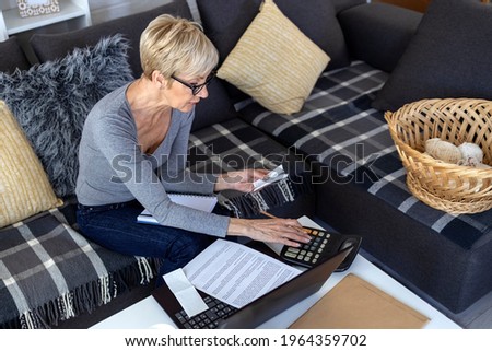 An older woman with short blonde hair sits on sofa in living room and works on laptop