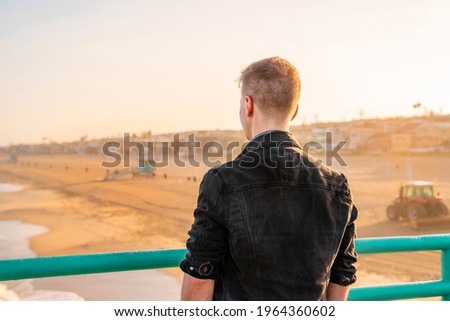 A young blond man in a denim jacket stands on a pier overlooking the beach in the early morning, Los Angeles, California