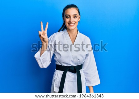 Beautiful brunette young woman wearing karate fighter uniform with black belt showing and pointing up with fingers number two while smiling confident and happy. 