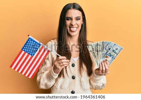 Beautiful brunette young woman holding united states flag and dollars sticking tongue out happy with funny expression. 