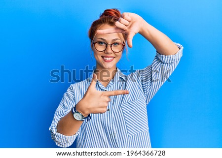 Young beautiful redhead woman wearing casual clothes and glasses over blue background smiling making frame with hands and fingers with happy face. creativity and photography concept. 