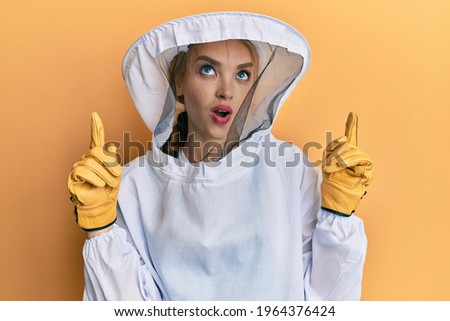 Beautiful blonde caucasian woman wearing protective beekeeper uniform amazed and surprised looking up and pointing with fingers and raised arms. 