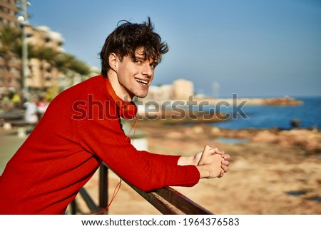 Young hispanic man using headphones leaning on the balustrade at the beach