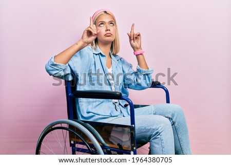 Beautiful blonde woman sitting on wheelchair pointing up looking sad and upset, indicating direction with fingers, unhappy and depressed. 