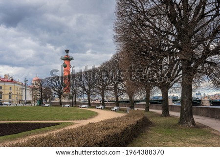 Russia, St. Petersburg, Vasilievsky Island. Famous landmark "Rostral Columns". Landscape park surrounded them. Cloudy spring day. Rows of pruned bare trees against backdrop of gray clouds.