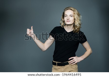 Young guy with long blond hair gesture catches a car hitchhiking or thumbs up. Isolated on gray background.