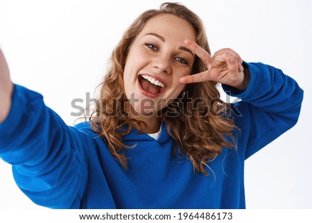 Positive attractive girl taking selfie, showing v-sign peace gesture near eye and holding camera with stretched hand, making photo on smartphone, white background
