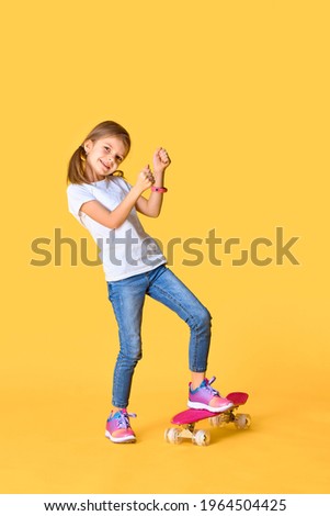 Stylish funny girl wearing white t-shirt, blue jeans and sneakers, standing on skateboard over yellow background.