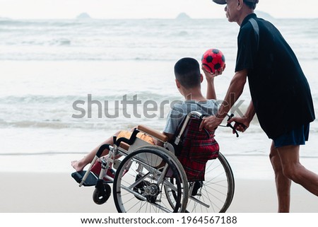 Asian special child on wheelchair holding ball, playing and exercise activity on sea beach at rainy day,Lifestyle of disability child, Life in the education age, Happy disabled kid in travel concept.