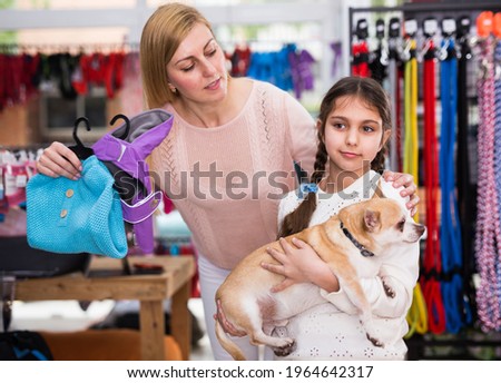 Cute girl with her positive mother and dog in pet shop looking through different clothes and accessories