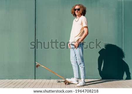 horizontal portrait of a caucasian young skateboarder looking at camera. He is wearing casual clothes and sunglasses. Green wall background