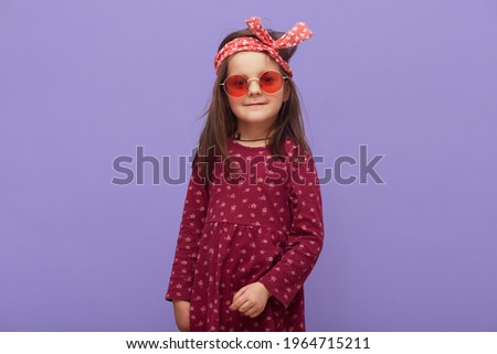 Charming little hipster fashionable girl dressed in burgundy dress, red headband and stylish glasses looking at camera, posing isolated on a purple background