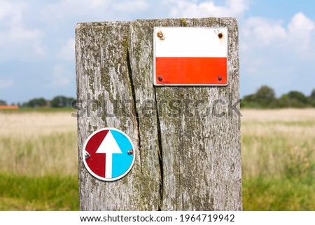 Two marking symbols in The Netherlands for European long distance hiking routes and Frisian hiking routes on a weathered wooden pole in front of a sunny landscape. Royalty-Free Stock Photo #1964719942
