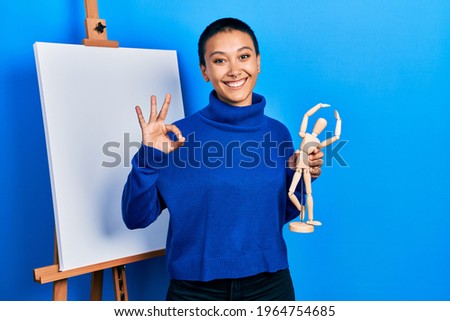 Beautiful hispanic woman with short hair holding manikin close to easel stand doing ok sign with fingers, smiling friendly gesturing excellent symbol 