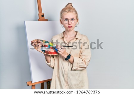 Beautiful caucasian woman with blond hair standing drawing with palette by painter easel stand depressed and worry for distress, crying angry and afraid. sad expression. 