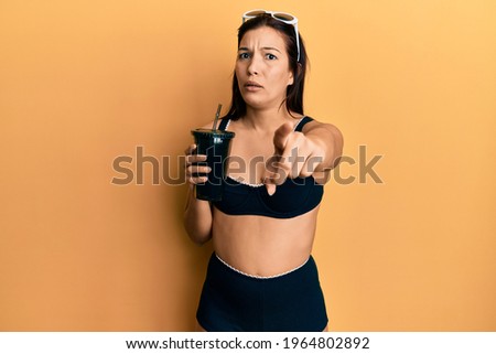 Young latin woman wearing bikini drinking soda pointing with finger to the camera and to you, confident gesture looking serious 