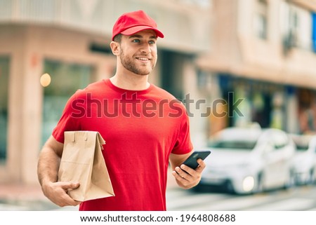Young caucasian deliveryman using smartphone and holding delivery paper bag at the city.