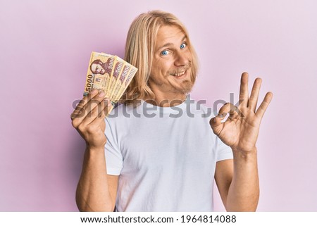 Caucasian young man with long hair holding 5000 hungarian forint banknotes doing ok sign with fingers, smiling friendly gesturing excellent symbol 
