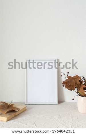 Home decor mocap, empty picture frame near white painted concrete wall, branches with dry leaves in a vase and old books with postcards