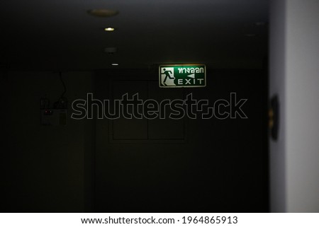 Green emergency exit sign in corridor point way out of resident apartment door way in Thai text information on dark background. Selective focus.