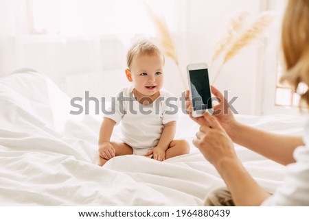 Cute baby is sitting on the bed and posing for his mom, mom is taking pictures of her baby on her mobile phone. Modern technologies, space for text