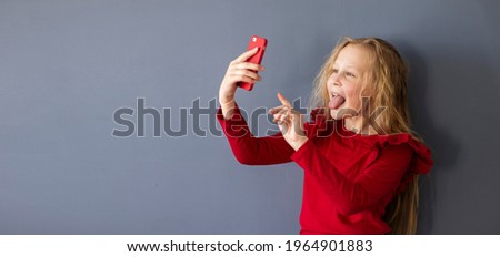 A contented blonde girl enjoys a message on a mobile phone, enjoys online communication, wears a red dress, isolated on a blue wall. Place for text Technology concept, selective focus