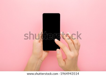 Young adult woman hand holding smartphone and touching black screen with finger on light pink table background. Pastel color. Empty place for text. Closeup. Point of view shot.
