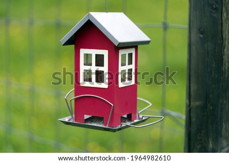Bird feeder in shape of a litte red house attached at wooden pole with meadow in the background.