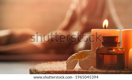 Warm inviting picture of beautiful spa composition with frangipani flower, oil flask, and candles. Blurred figures of the masseur and his guest in background. Royalty-Free Stock Photo #1965045394