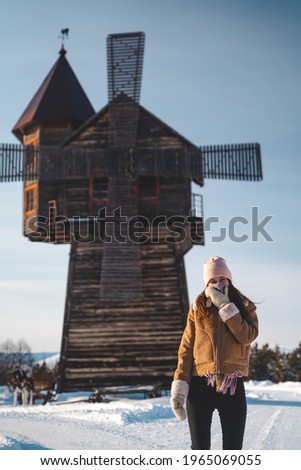 A young woman in warm clothes with a hat, a scarf, mittens, a sheepskin coat in frost stands on a snow-covered path against the background of an old mill in the reserve